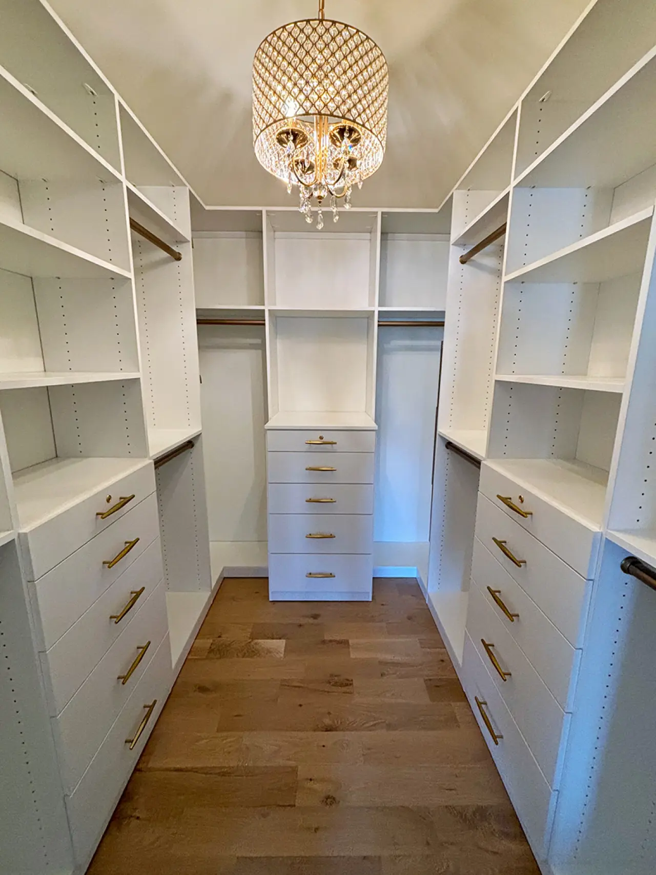 White walk-in closet with shelves, drawers, and gold handles, under a decorative chandelier.