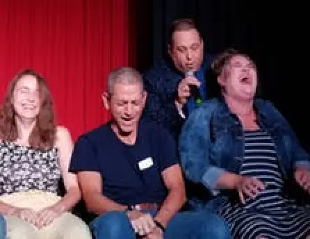 Three adults sit laughing on stage as a man with a microphone stands behind them performing.