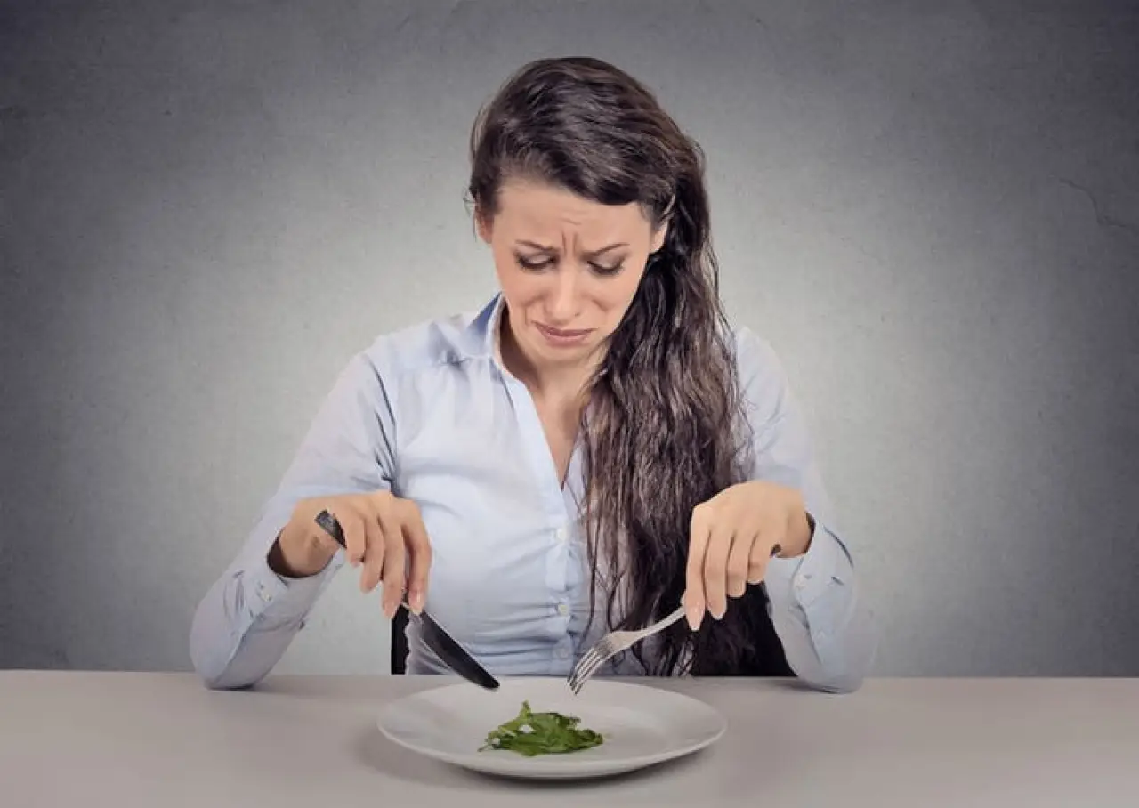 Woman frowning at a small portion of greens on a plate, holding a fork and knife.
