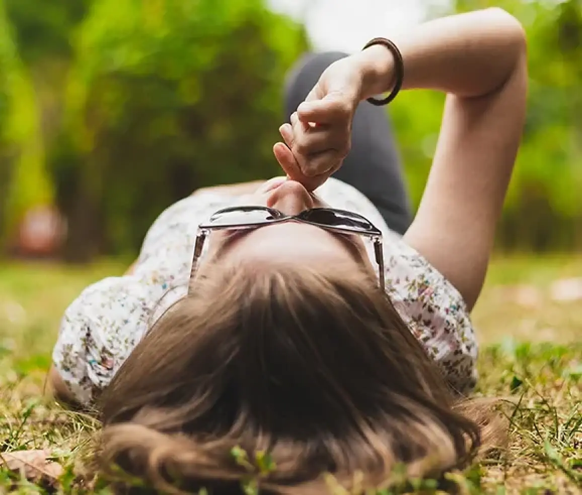Person lying on grass outdoors, wearing sunglasses and floral top, with hand near face.
