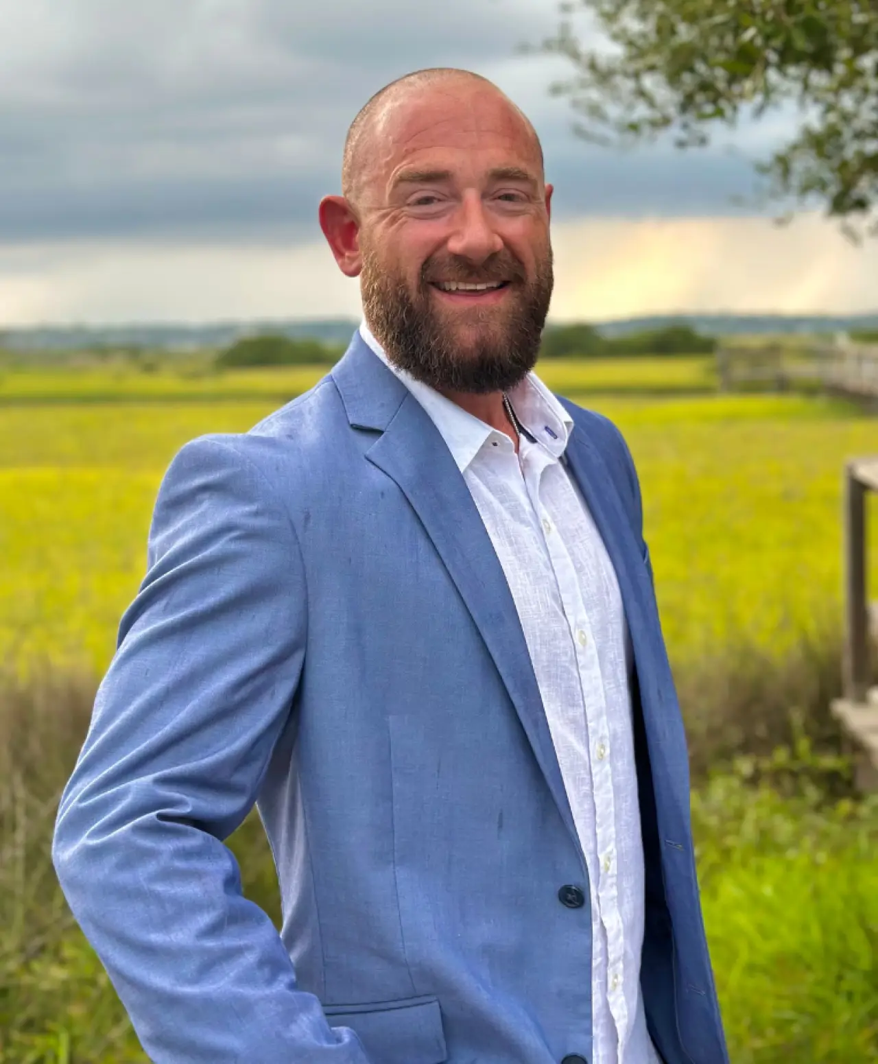 A man in a light blue suit jacket smiles outdoors with green fields in the background.