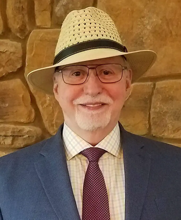 Man in a suit, wearing a straw hat and glasses, smiles against a stone wall background.