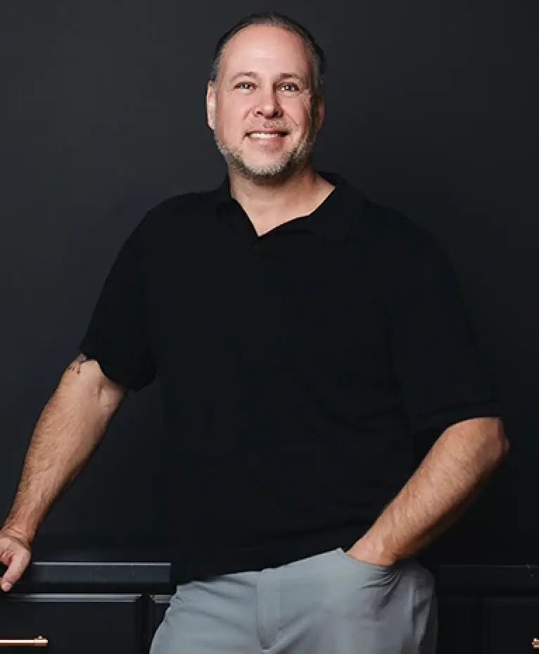 Smiling man in a black polo shirt and gray pants stands against a dark background.