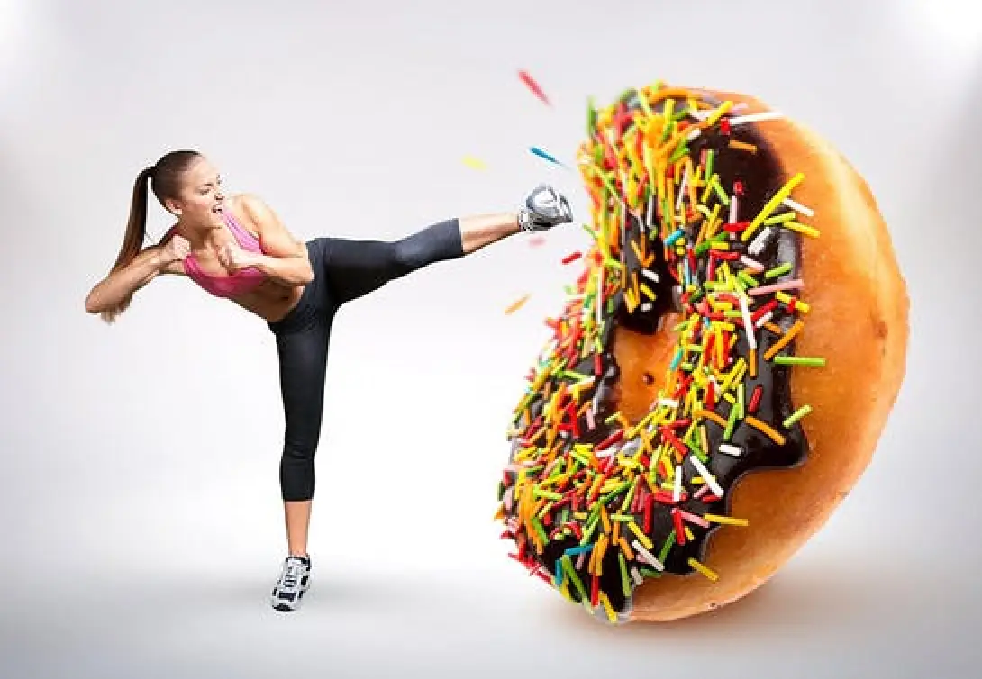 Woman in athletic wear kicks a large donut covered in chocolate and sprinkles.