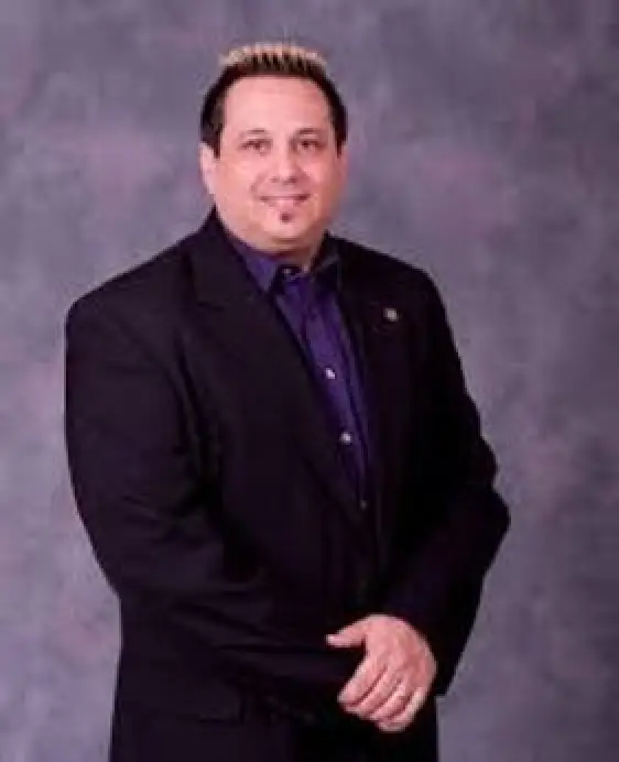 Man in a black suit with short hair standing against a gray studio background, smiling slightly.