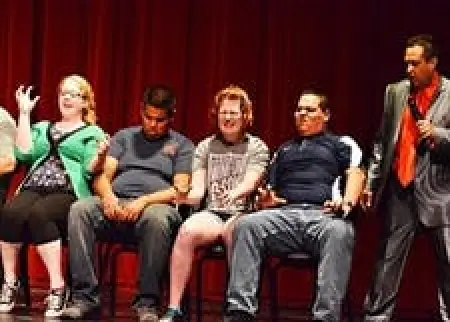 Five people sit on stage, four in chairs and one standing, with a red curtain as the backdrop.