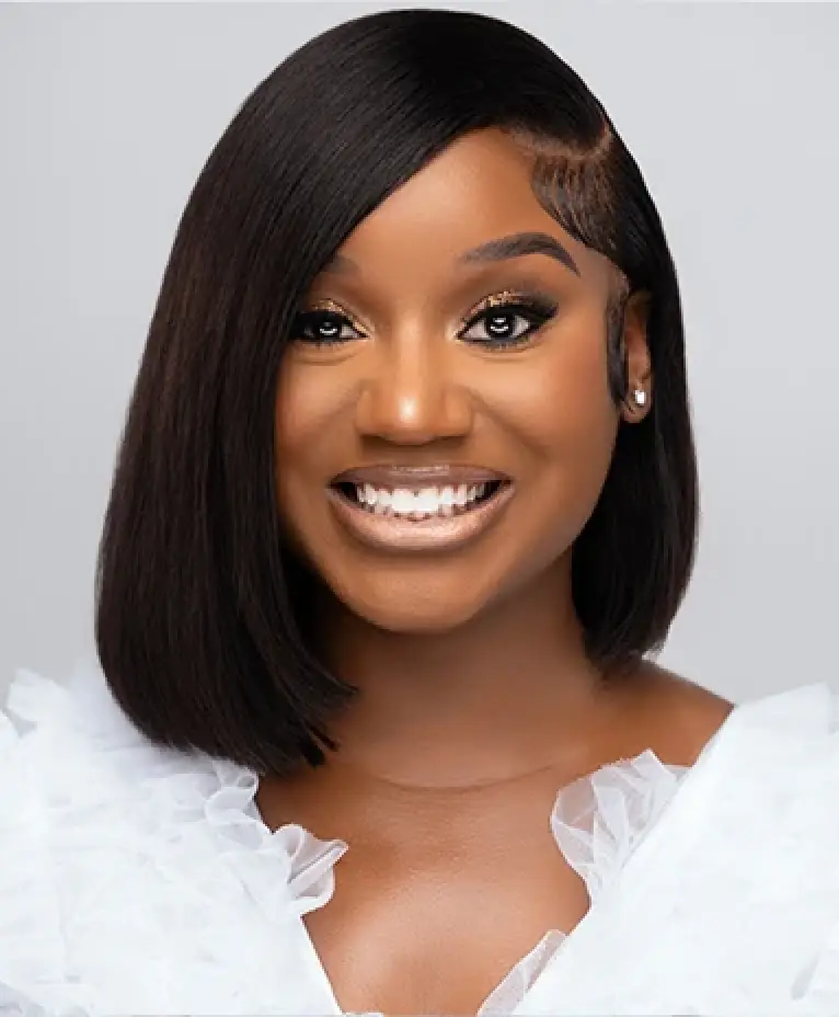 Smiling woman with straight, shoulder-length hair wearing a white ruffled top against a gray background.