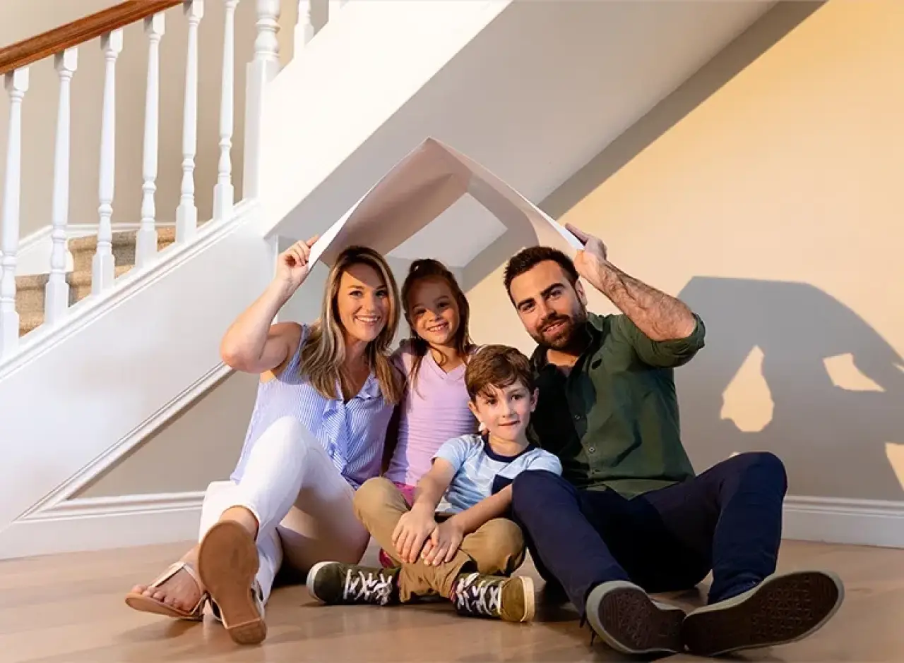 A happy family of four sits on the floor, holding a roof-shaped board over their heads.