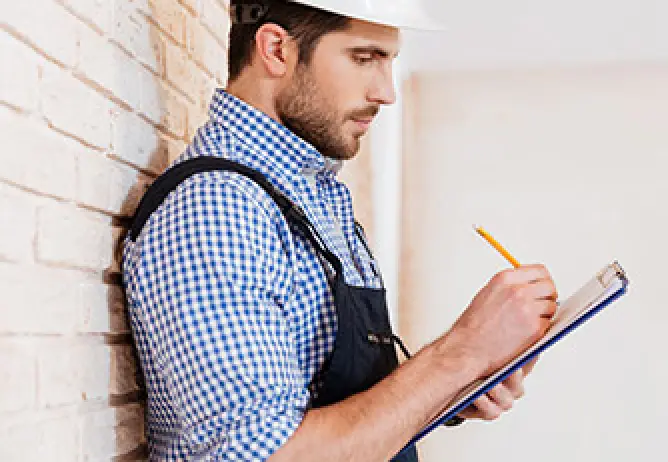 A man in a blue checkered shirt, black overalls, and a hard hat writing on a clipboard
