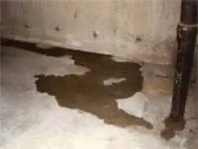 Water pooling on a concrete basement floor near a wall and metal pipe, possibly from a leak.