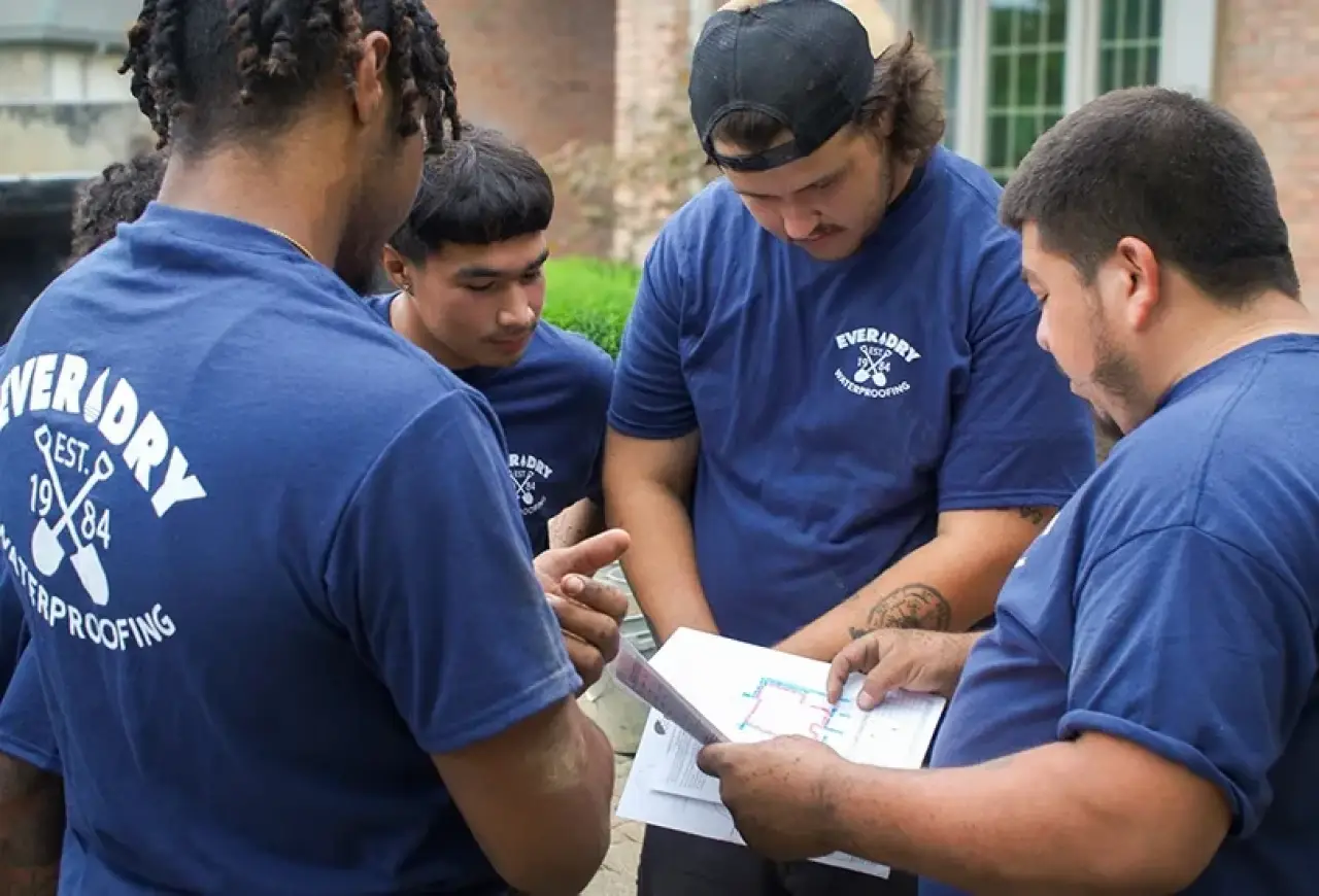 Four men in blue shirts review papers and plans together outside a brick building.