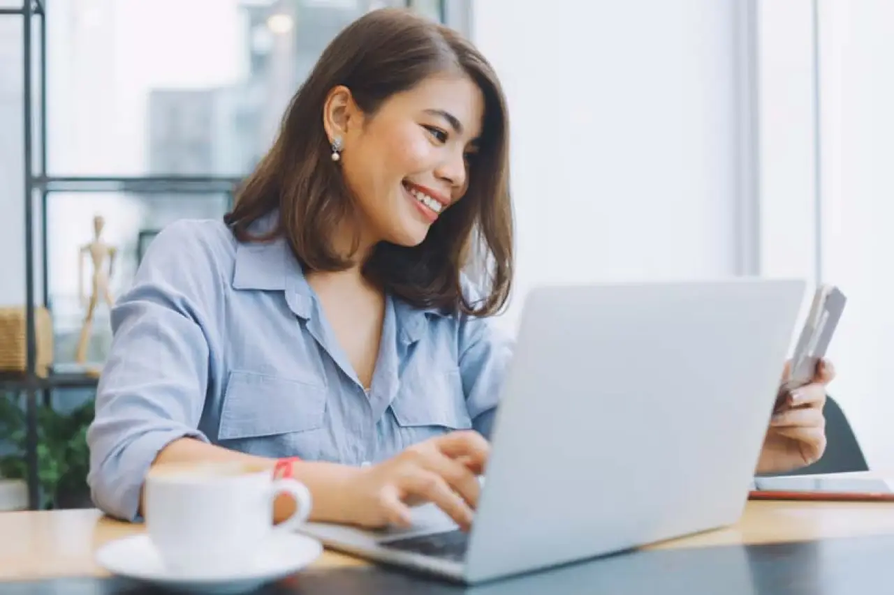 Smiling woman using a laptop at a cafe, holding a smartphone, with a cup of coffee on the table.