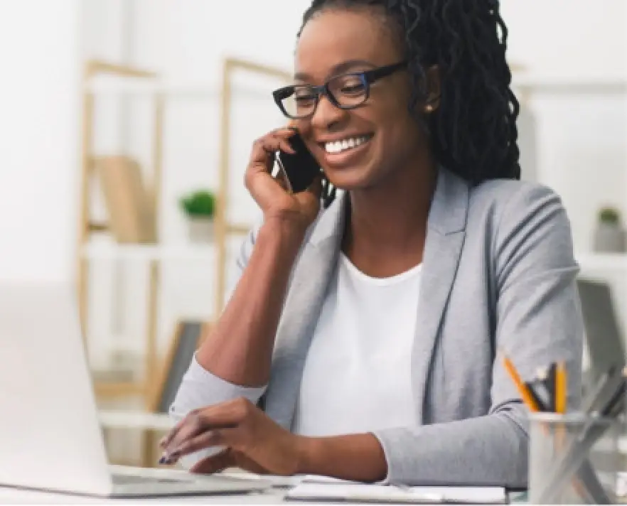 Smiling woman in glasses talking on phone and working on laptop at a desk.