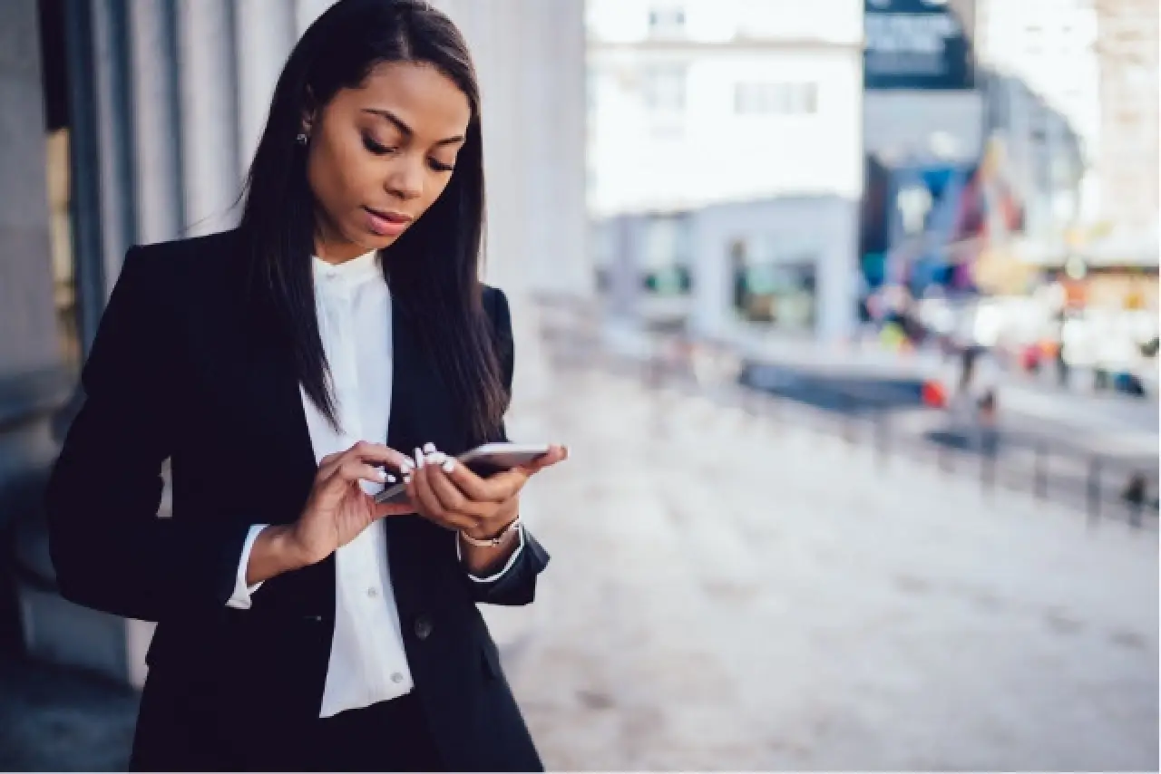 Woman in a black suit stands outside, looking down at her phone and typing.