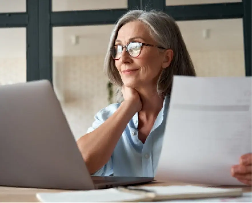 Smiling older woman with glasses looks at laptop, holding a paper in her hand at a desk.