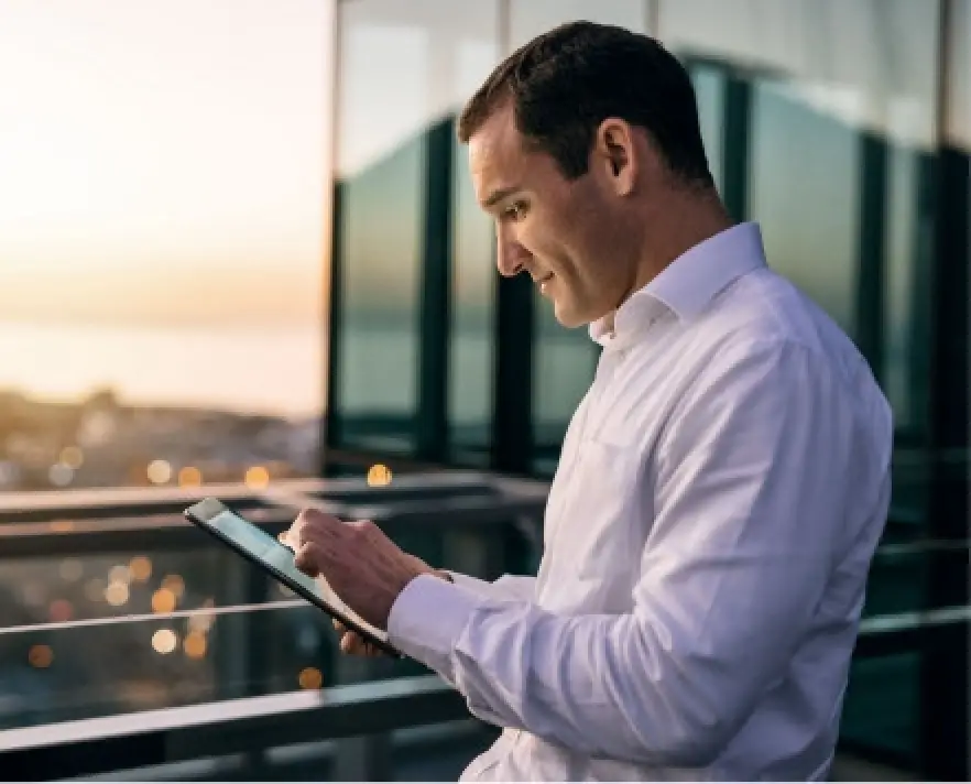 Man in white shirt using a tablet on a balcony at sunset with city lights in the background.