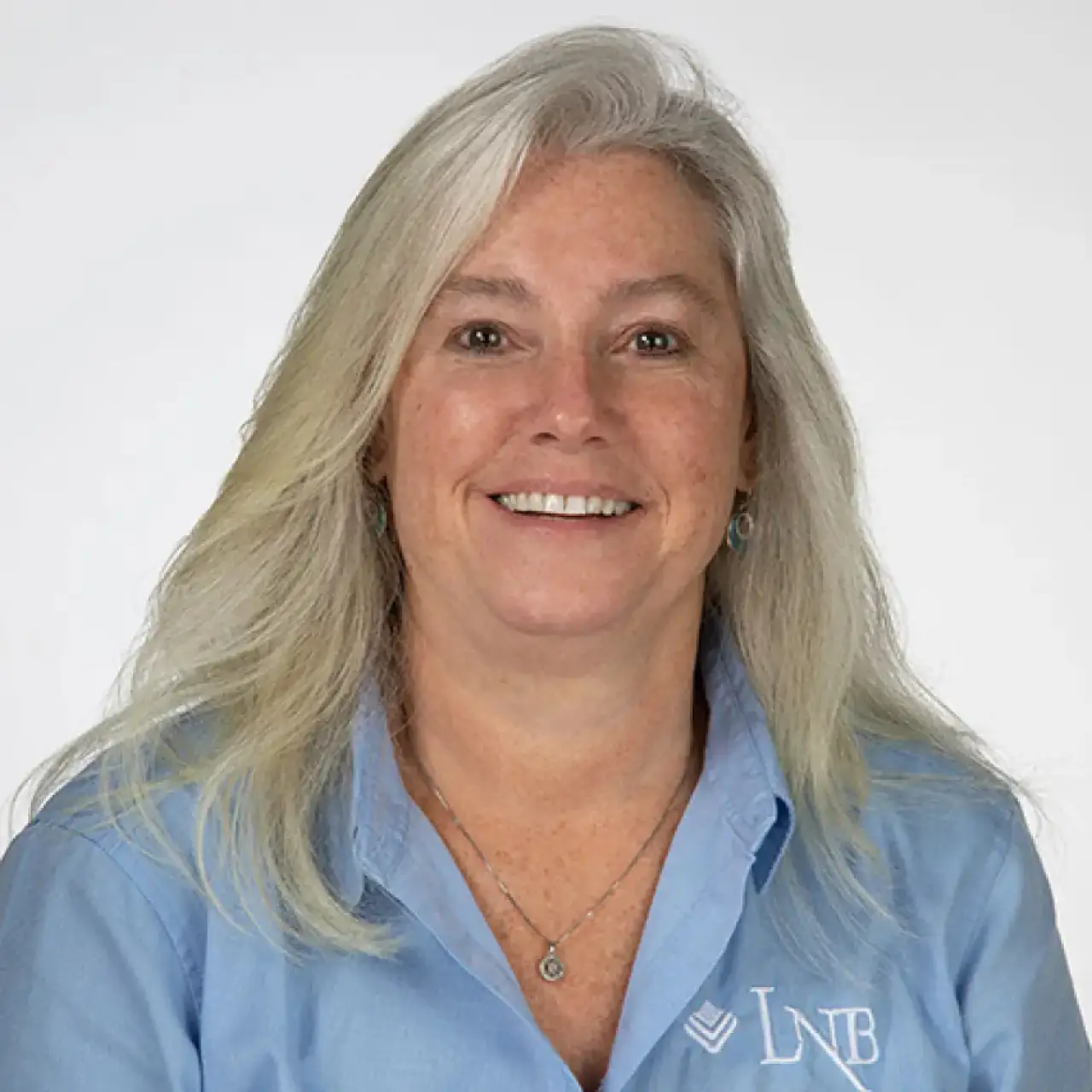 Smiling woman with long gray hair wearing a light blue LNB shirt against a white background.
