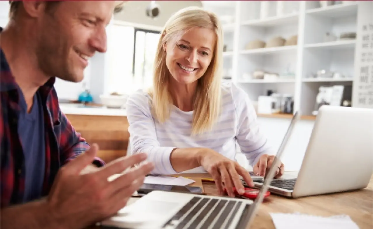 Smiling couple works together on laptops at a kitchen table with documents and coffee.