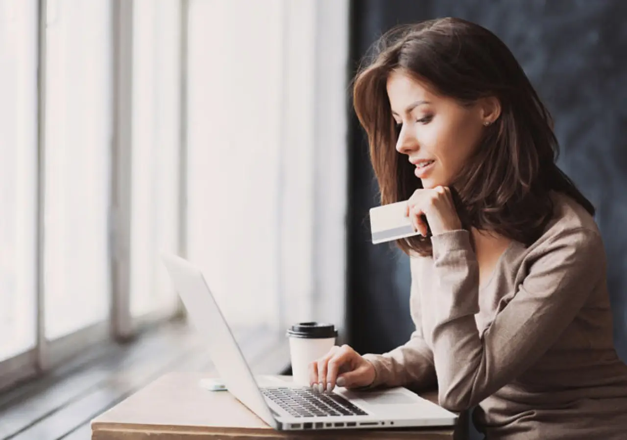 Woman holding a credit card and using a laptop at a desk with a coffee cup nearby.