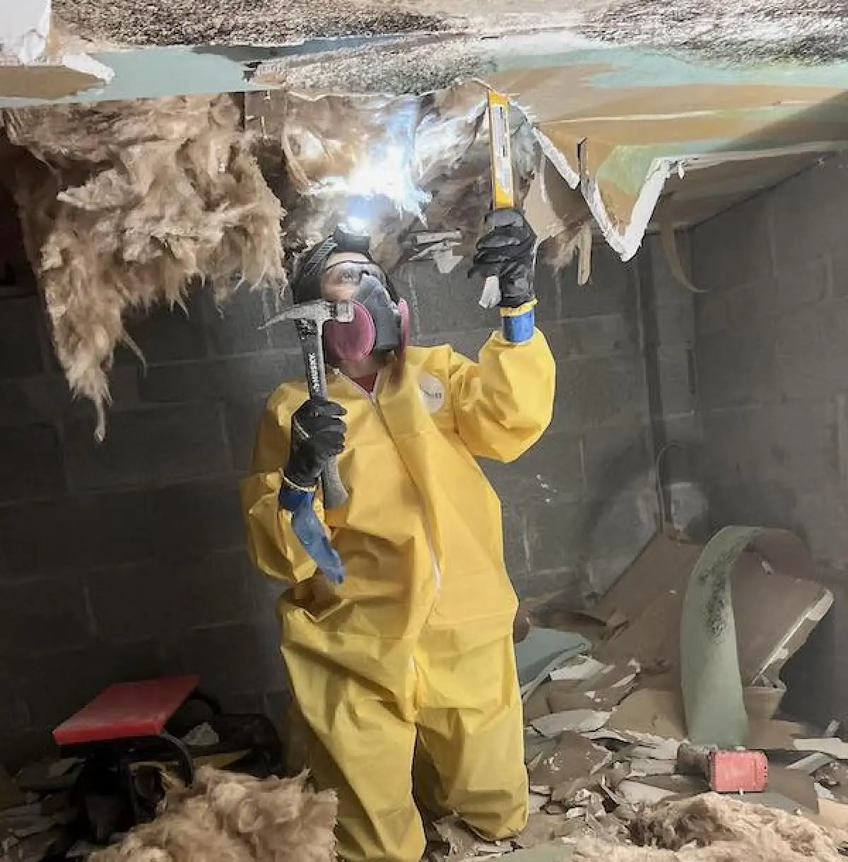 Person in yellow protective suit tearing down ceiling insulation and debris in a basement.