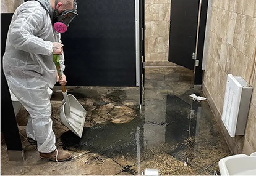 Person in protective gear cleaning a flooded restroom with a shovel.