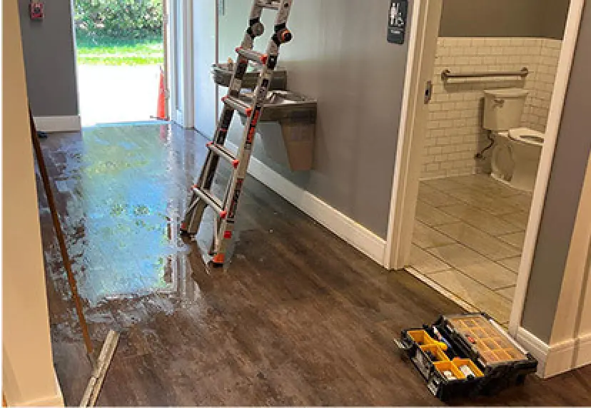 A ladder and toolbox sit near a wet floor outside a restroom with an accessible toilet.