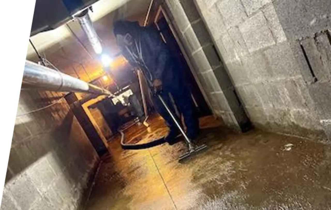 Person cleaning a flooded basement with a vacuum hose, dimly lit environment.