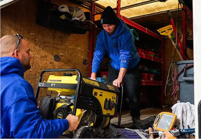 Two people in a workshop loading a yellow generator into a vehicle.