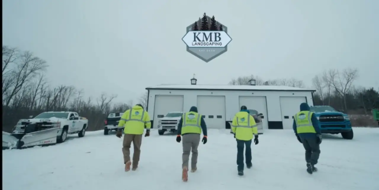 Four workers in high-visibility jackets walk toward a snowy garage with KMB Landscaping logo above.