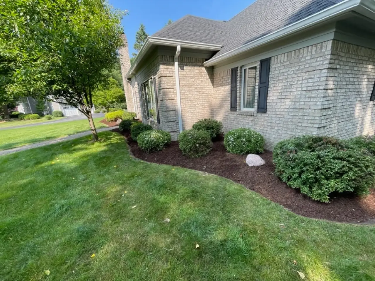 Brick house with black shutters, shrubs, and fresh mulch along a green, well-kept lawn.