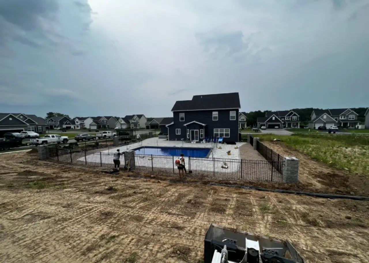 A fenced backyard pool behind a dark house, with workers finishing landscaping nearby.