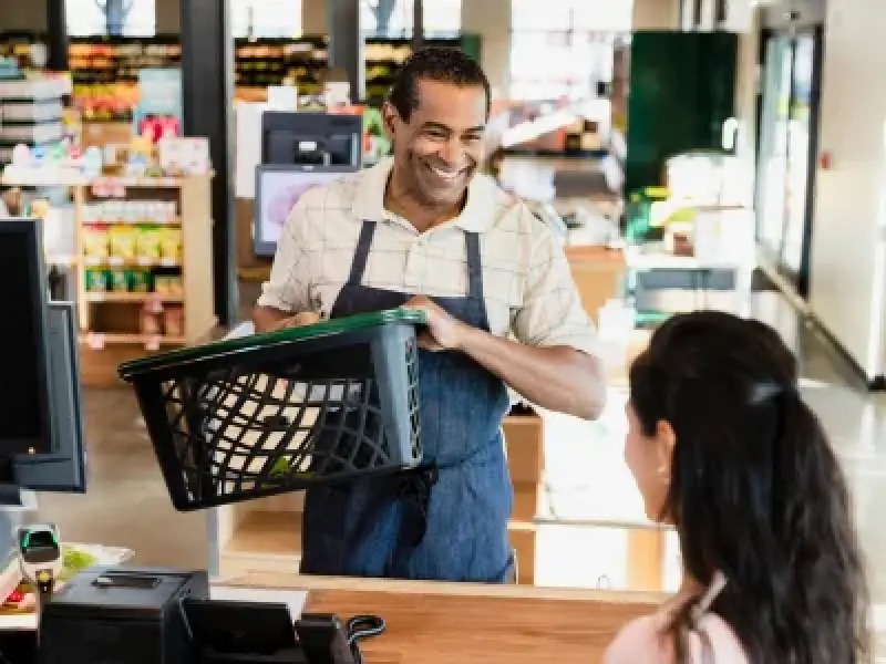 Smiling cashier hands a shopping basket to a customer at a grocery store checkout counter.