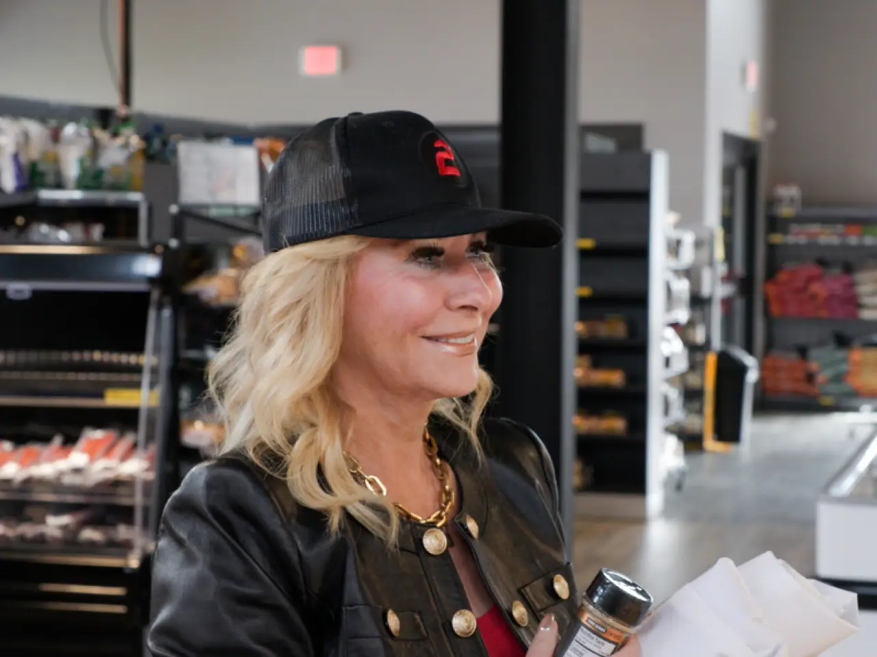 Smiling woman in a black cap and leather jacket holds items in a brightly-lit store.