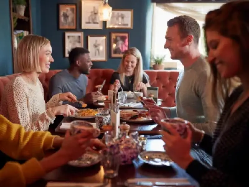 Six friends smile and enjoy a meal together at a restaurant, seated around a table.