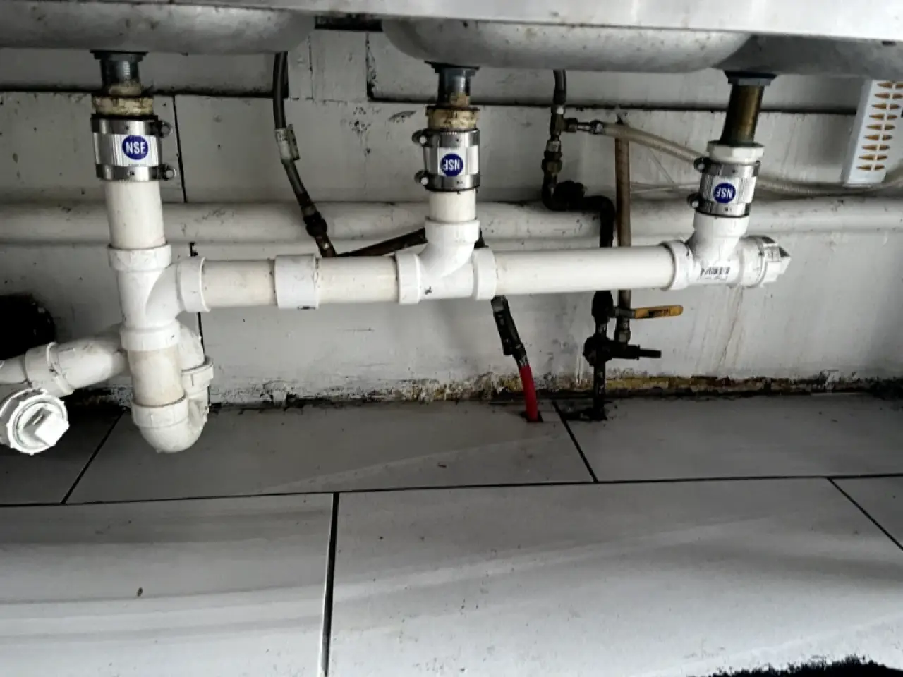 Grimy plumbing and tile beneath a commercial sink.