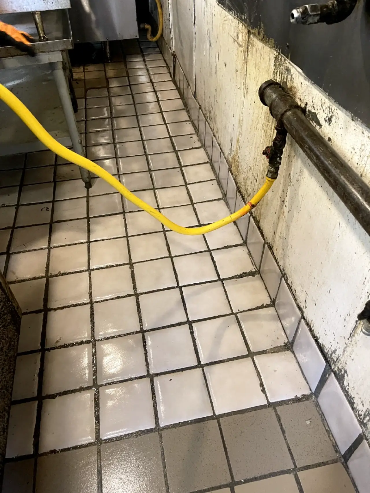 Grease-stained commercial kitchen wall beside a wet tiled floor and cleaning hose.