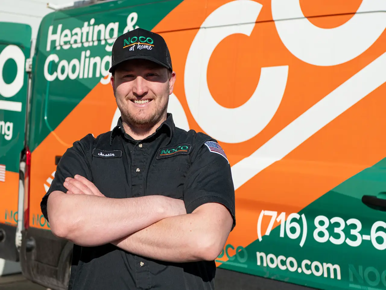 Smiling technician stands with arms crossed in front of a NOCO Heating Cooling service van.