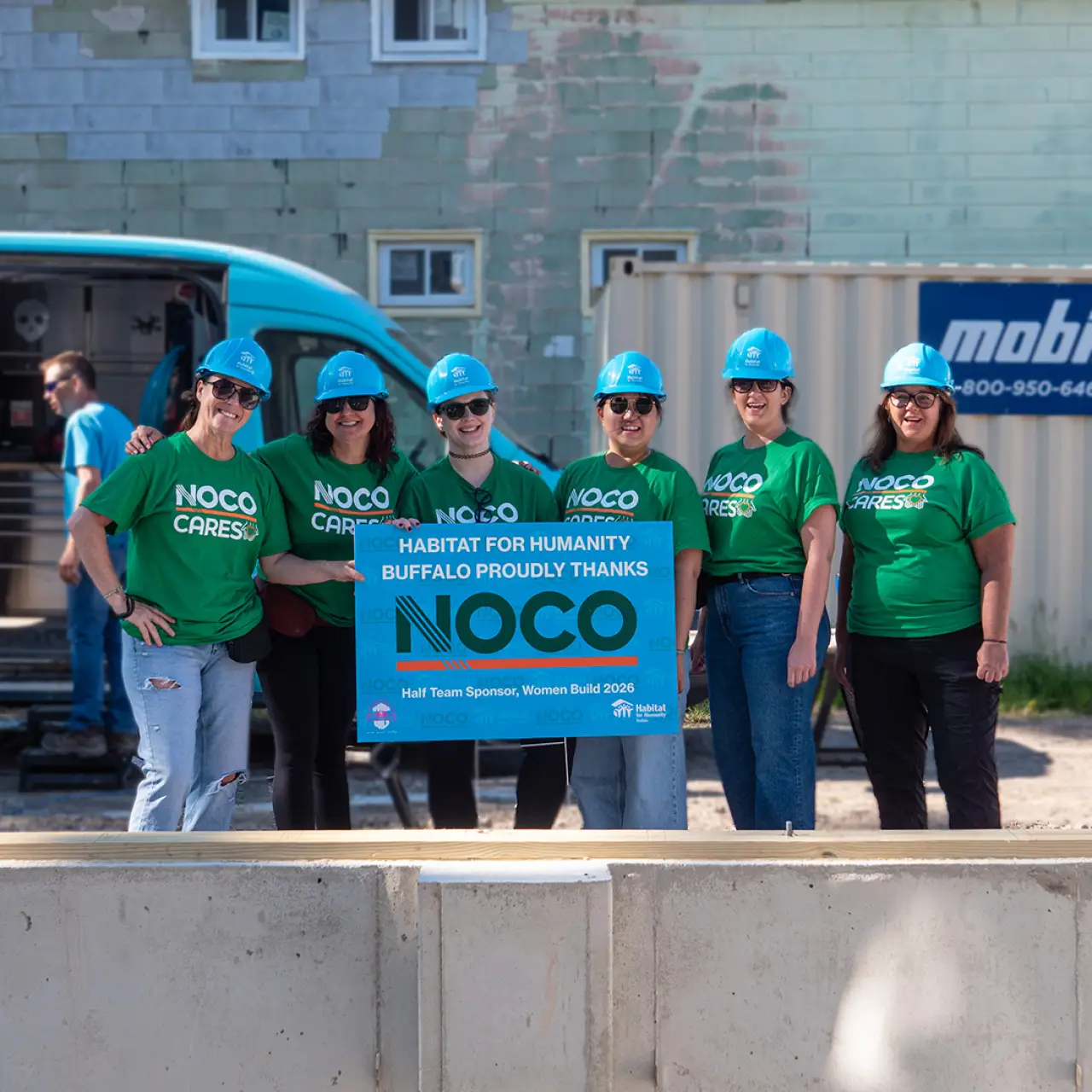 NOCO team members in NOCO Cares shirts and hard hats holding a Habitat for Humanity Buffalo thank-you sign at a Women Build site
