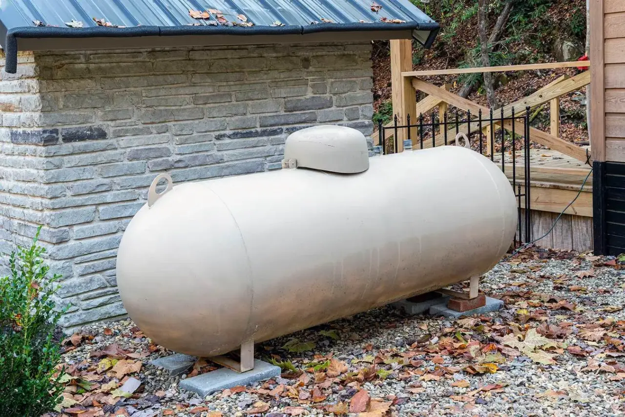 A beige propane tank sits outdoors on gravel near a house, with autumn leaves scattered around.