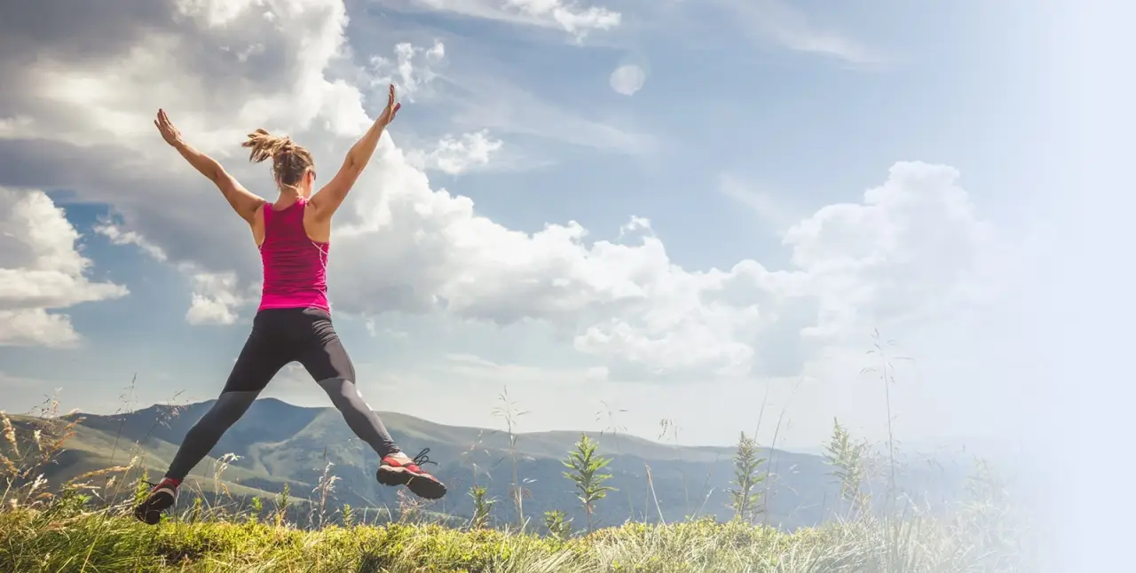 person in athletic wear jumping joyfully on a grassy hilltop with cloudy sky in the background