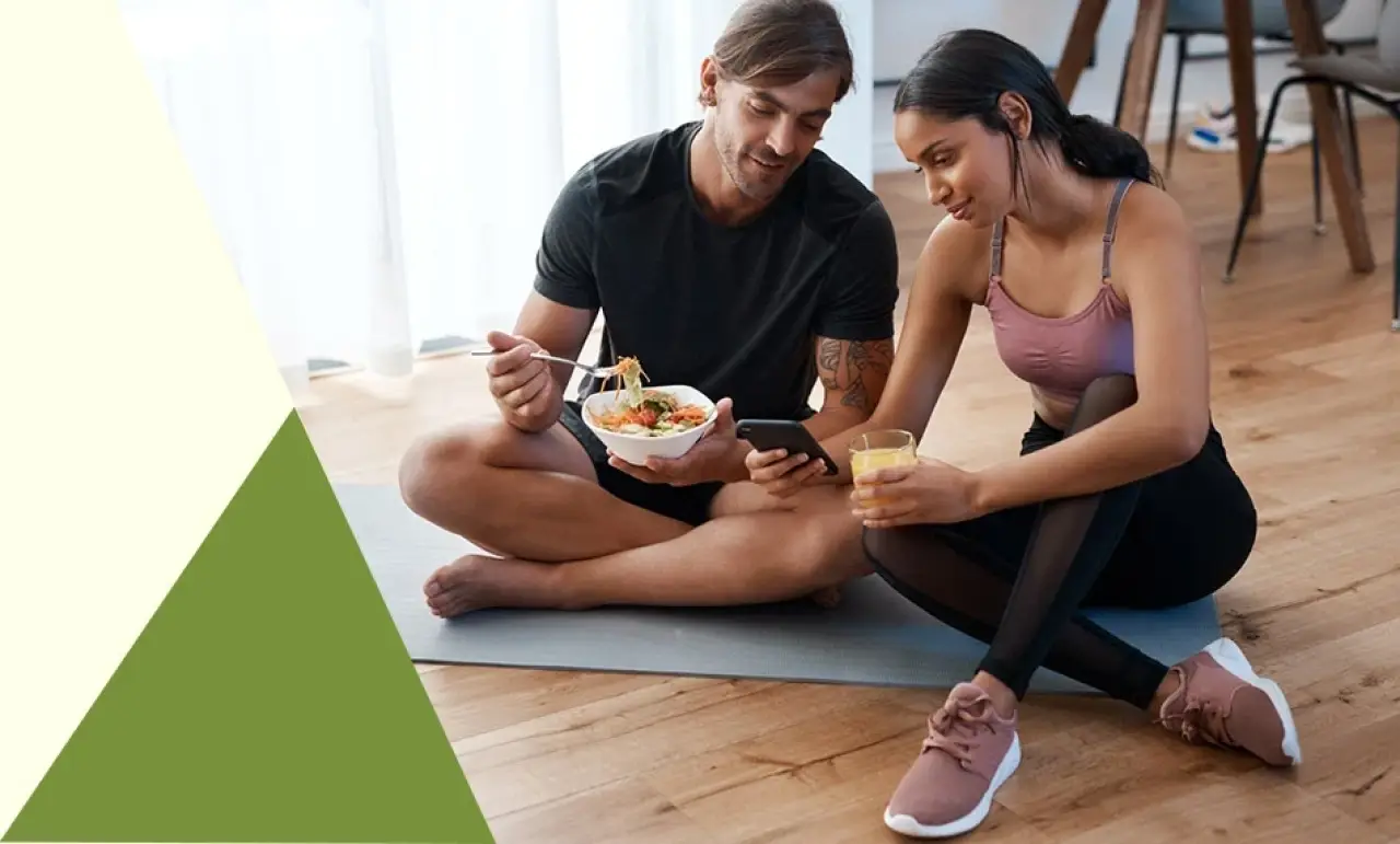 two people sit on a mat, eating and using a smartphone in a bright, casual setting