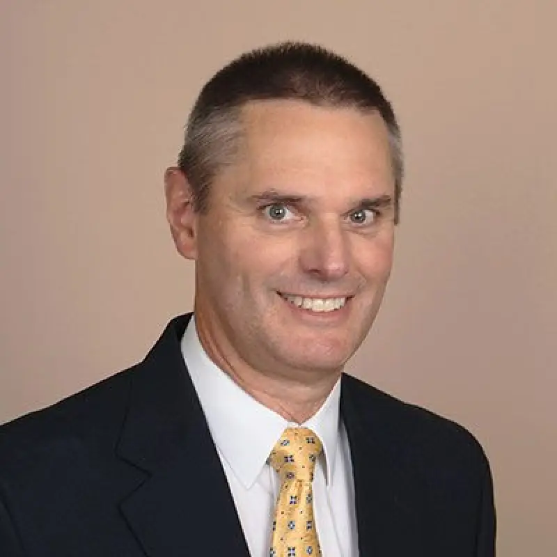 Smiling man in a suit and yellow tie poses against a plain, light-colored background.