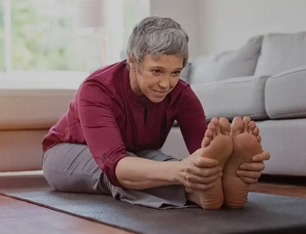 older adult stretches forward to touch their toes on a yoga mat in a living room