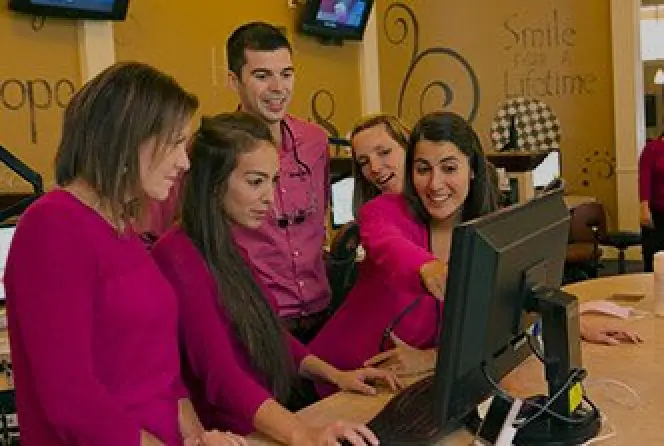 Four women and one man in magenta tops look at a computer screen and smile together.
