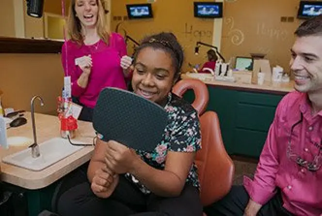 A smiling girl in a dental chair looks at her teeth in a mirror with two happy dentists nearby.
