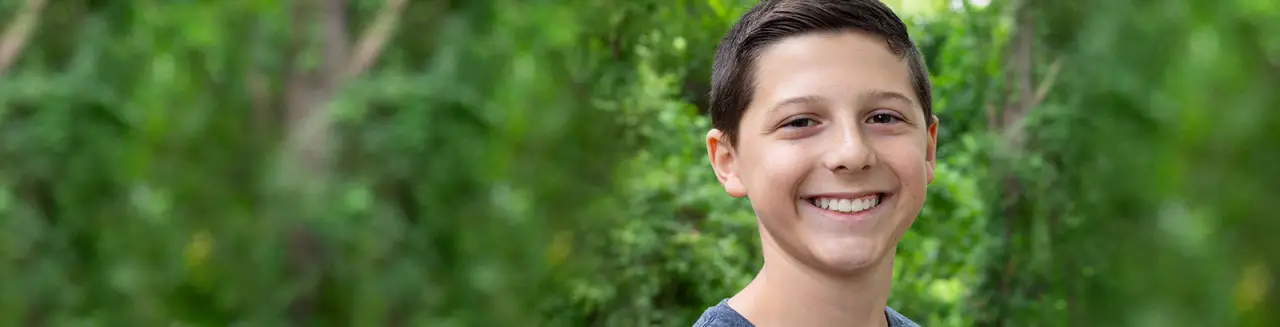 Smiling boy with short brown hair stands outdoors in front of blurred green trees.