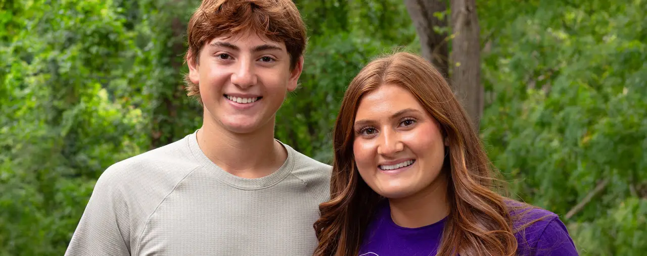 Two young adults smiling outdoors in front of green trees, one in gray and one in purple.
