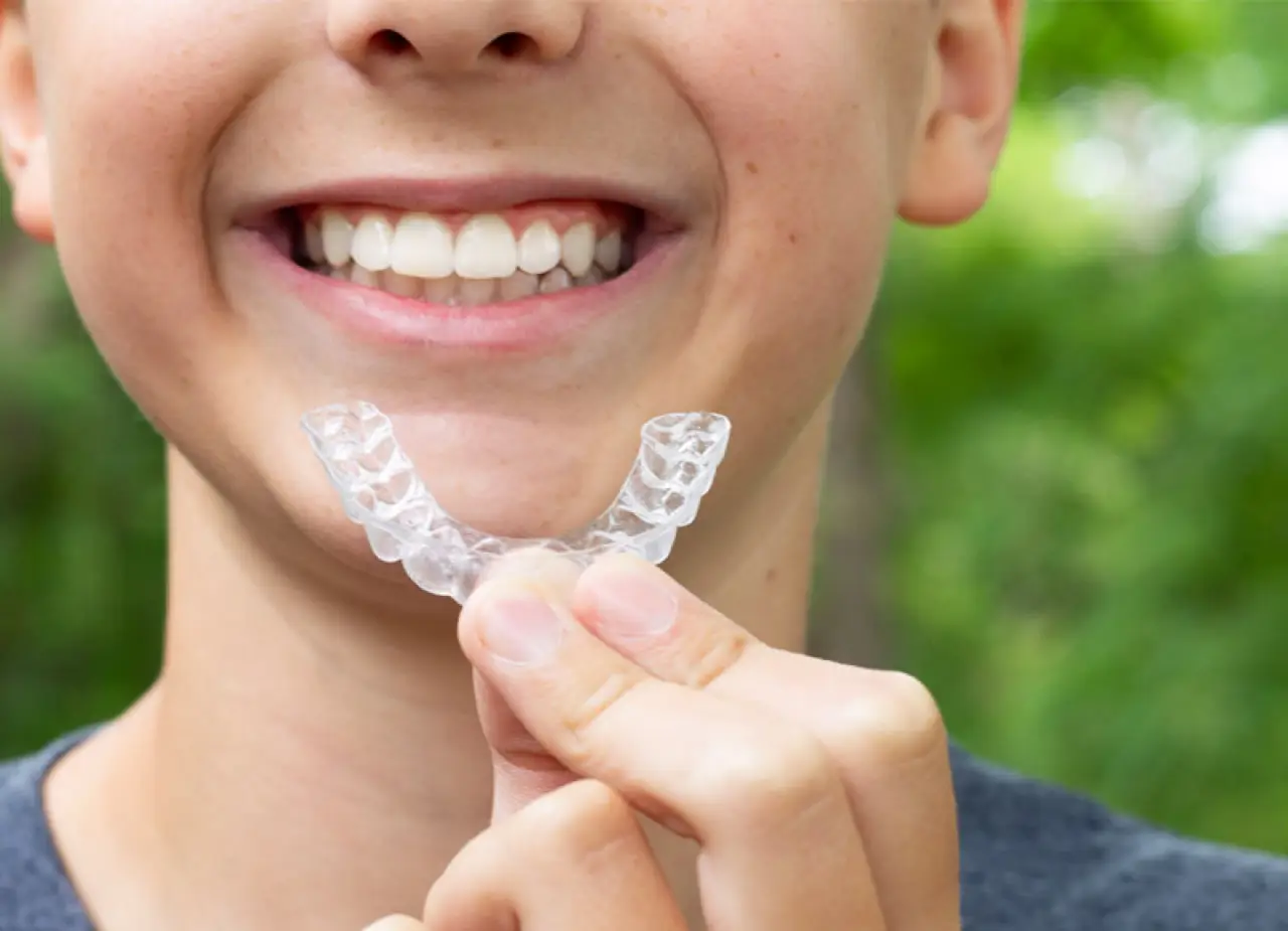 Smiling child holding a clear dental aligner in front of their teeth, outdoors.