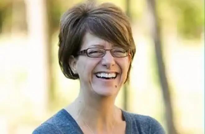 Woman with short brown hair and glasses smiling outdoors, wearing a gray top.