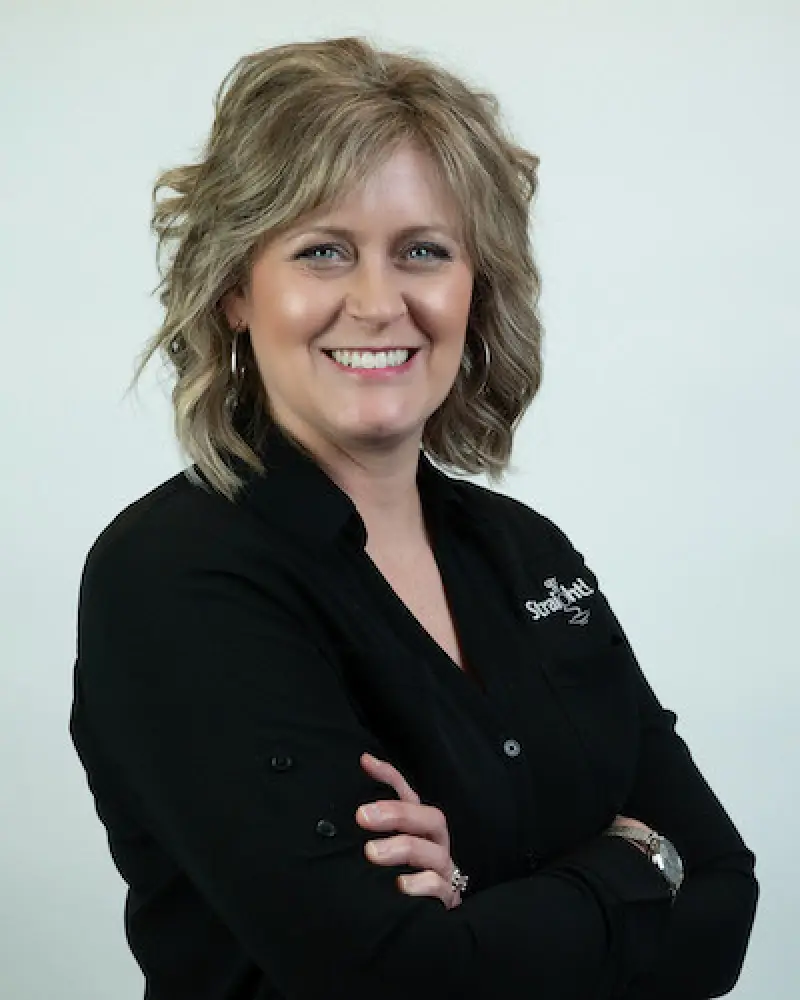 Smiling woman with wavy blond hair in a black shirt, standing with arms crossed.